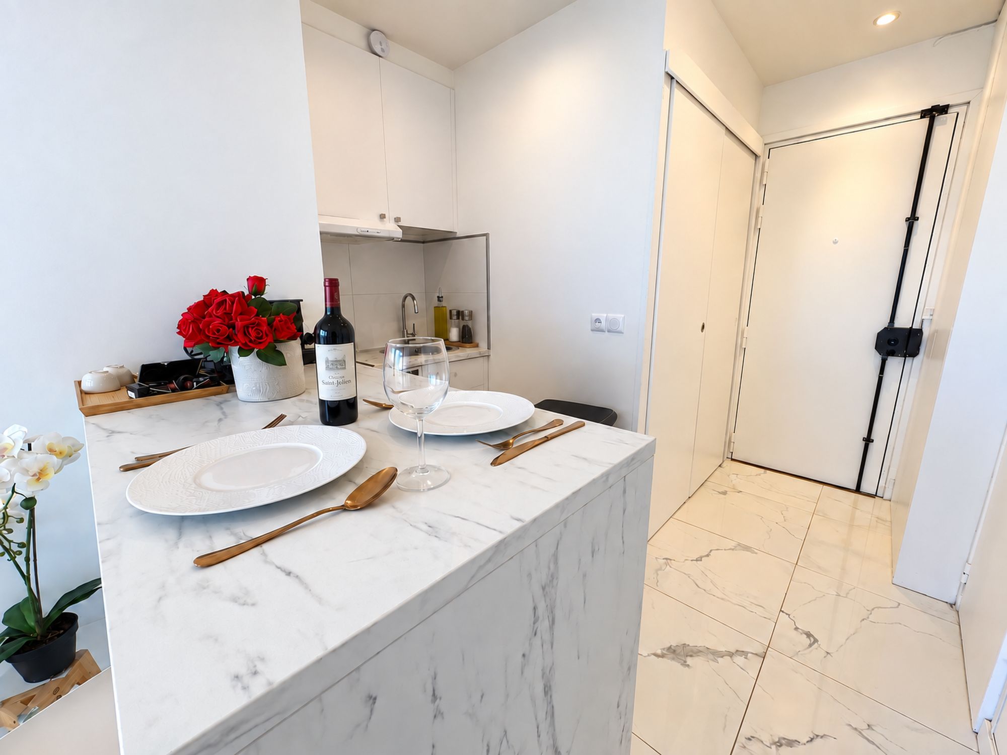 Une cuisine moderne avec des meubles blancs, un plan de travail en marbre et un coin repas agrémenté d’assiettes, de couverts et d’une bouteille de vin.