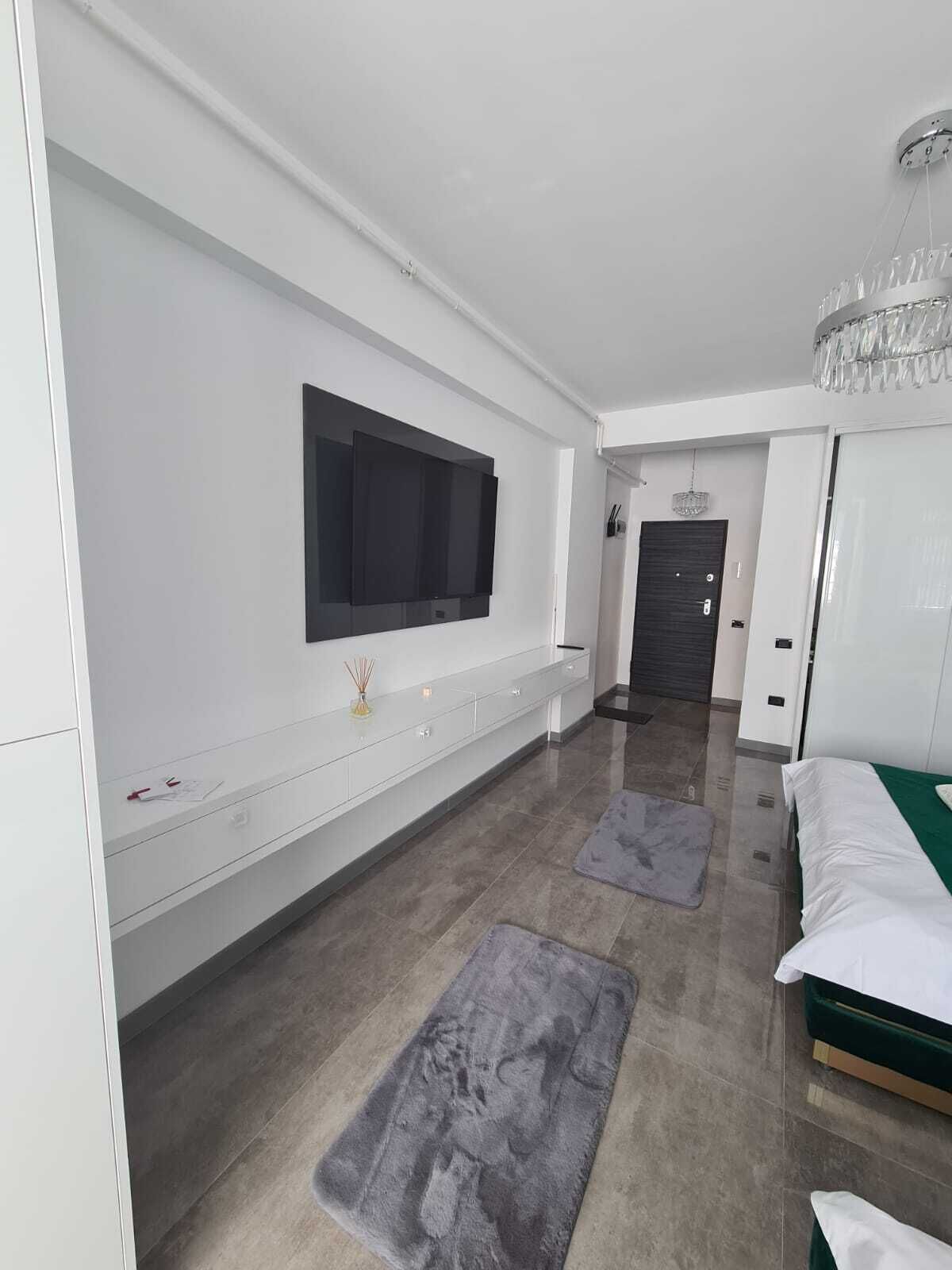 A modern hotel room with a large flat-screen TV mounted on the wall, a bed with a green bedspread, and a chandelier.