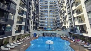 Un moderno edificio de apartamentos con una piscina central rodeada de sillones de descanso y una zona de juegos para niños.