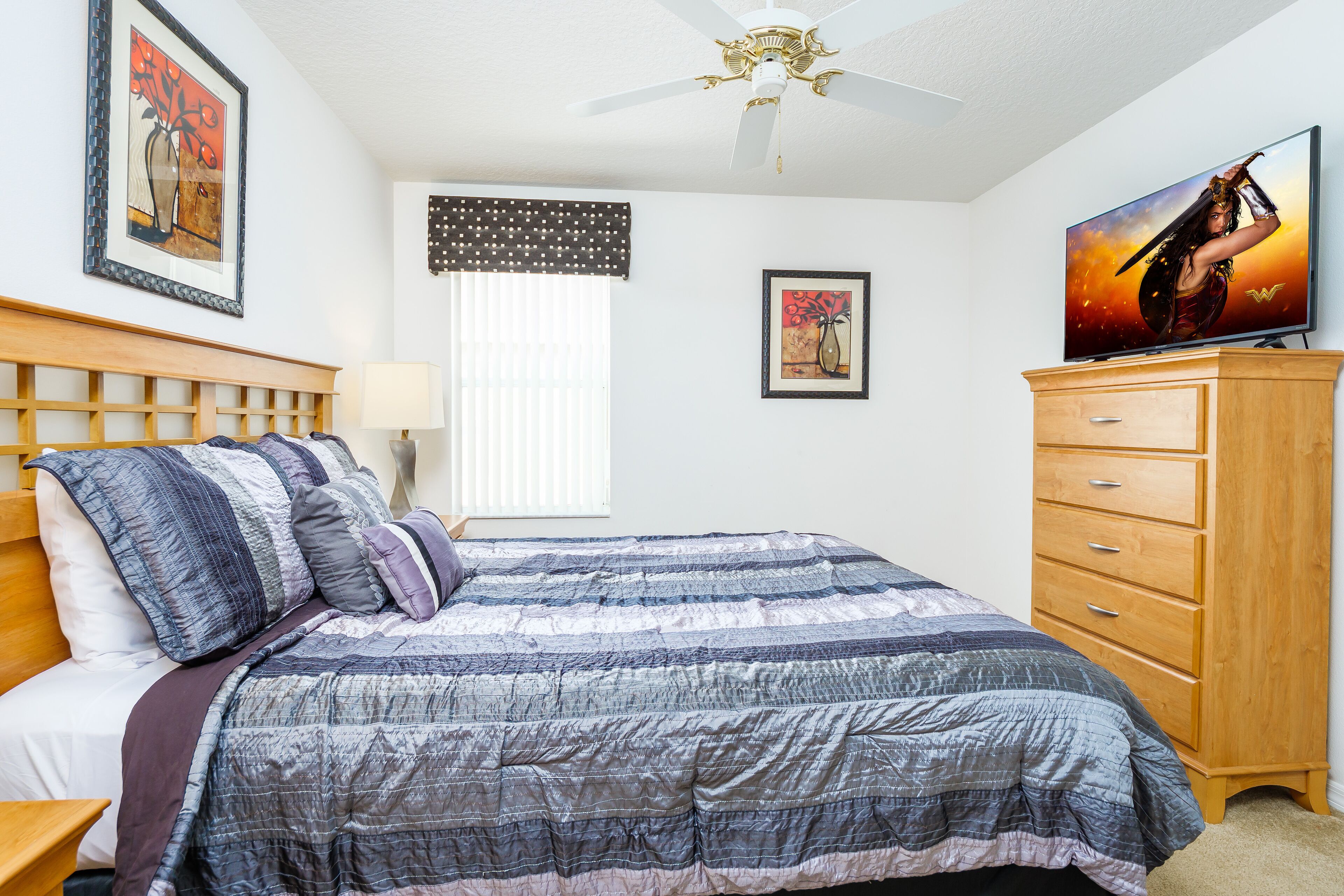 Un dormitorio con cama, cómoda de madera, ventilador de techo y televisor sobre un mueble de madera.