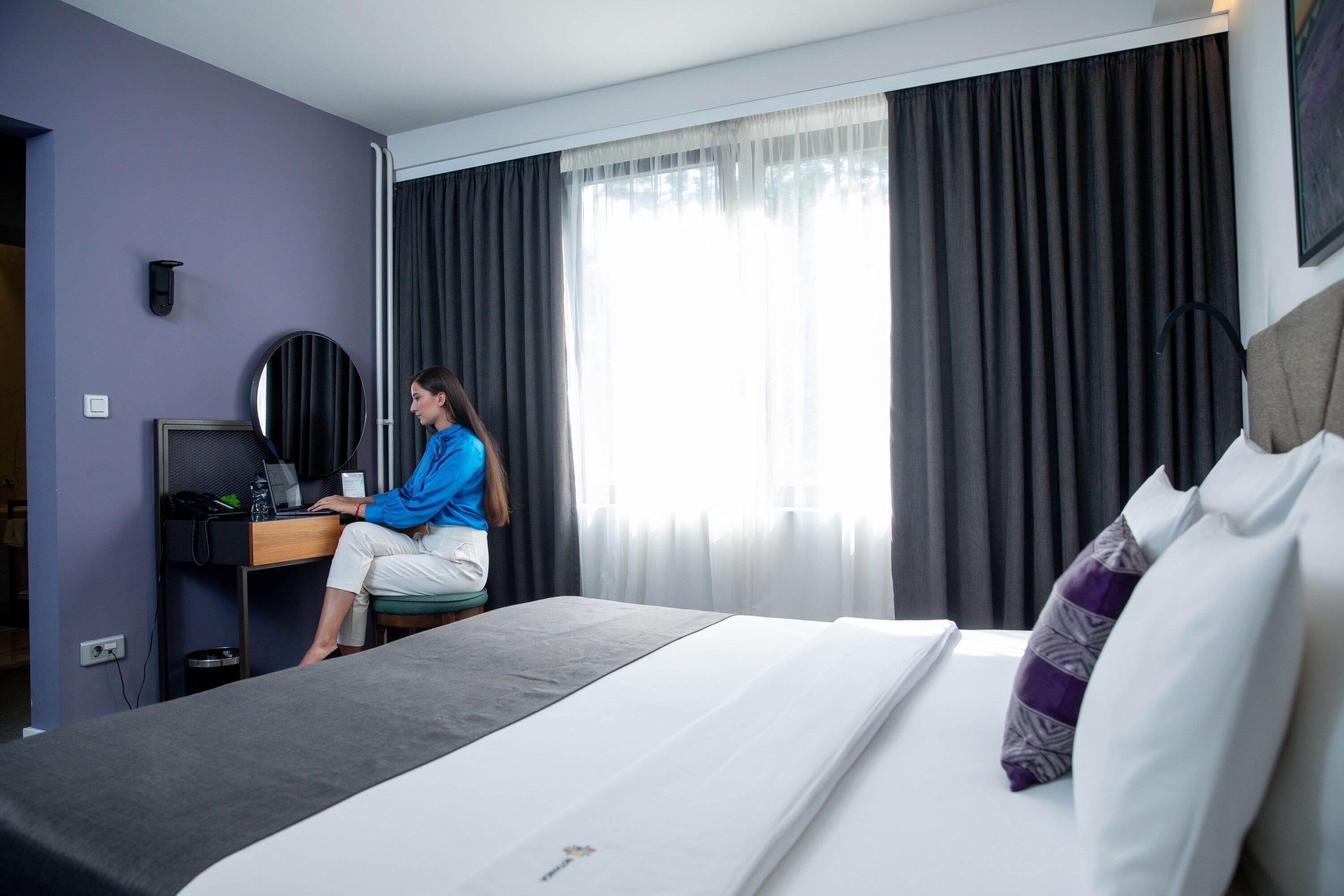 A hotel room with a bed, a desk, a chair, a computer, and a window with curtains.
