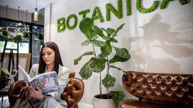 A woman reading a magazine in a room with a 'BOTANICA' sign, leather furniture, and potted plants. - Botanica Art (Belgrade)