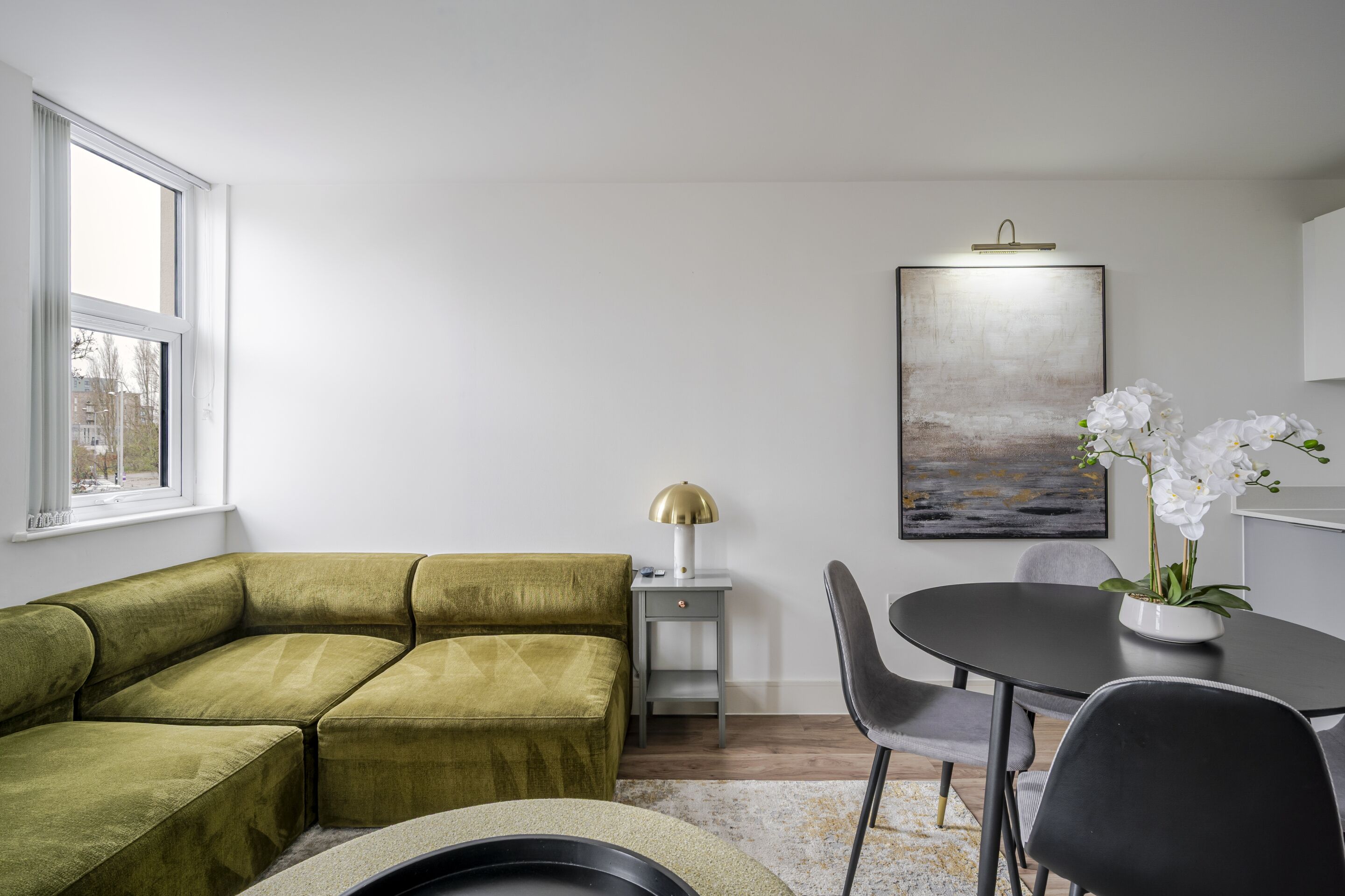 A modern living room with a green sectional sofa, a small side table with a lamp, a dining table with chairs, and a framed artwork on the wall.