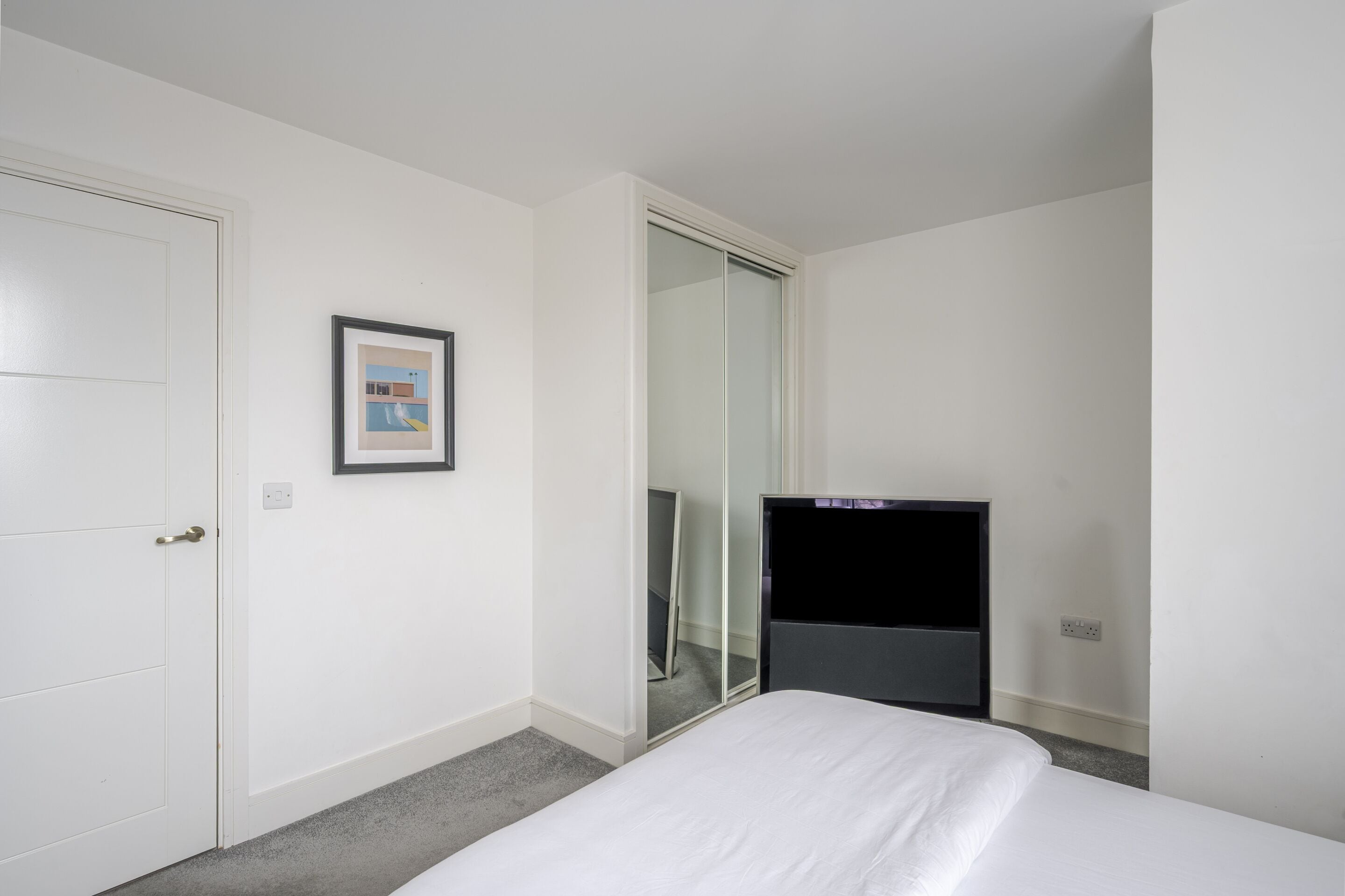 Una habitación de hotel con una cama, una puerta, un cuadro enmarcado, un espejo y un televisor.