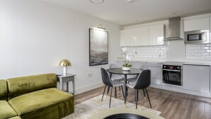 Cocina moderna con armarios blancos, un microondas y un horno. Hay un comedor con mesa y sillas, y un sofá verde en la sala de estar.
