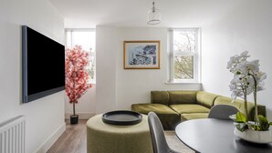 Una sala de estar moderna con un televisor de pantalla plana, un otoman redondo, un sofá verde, una mesa de comedor y una orquídea en maceta.