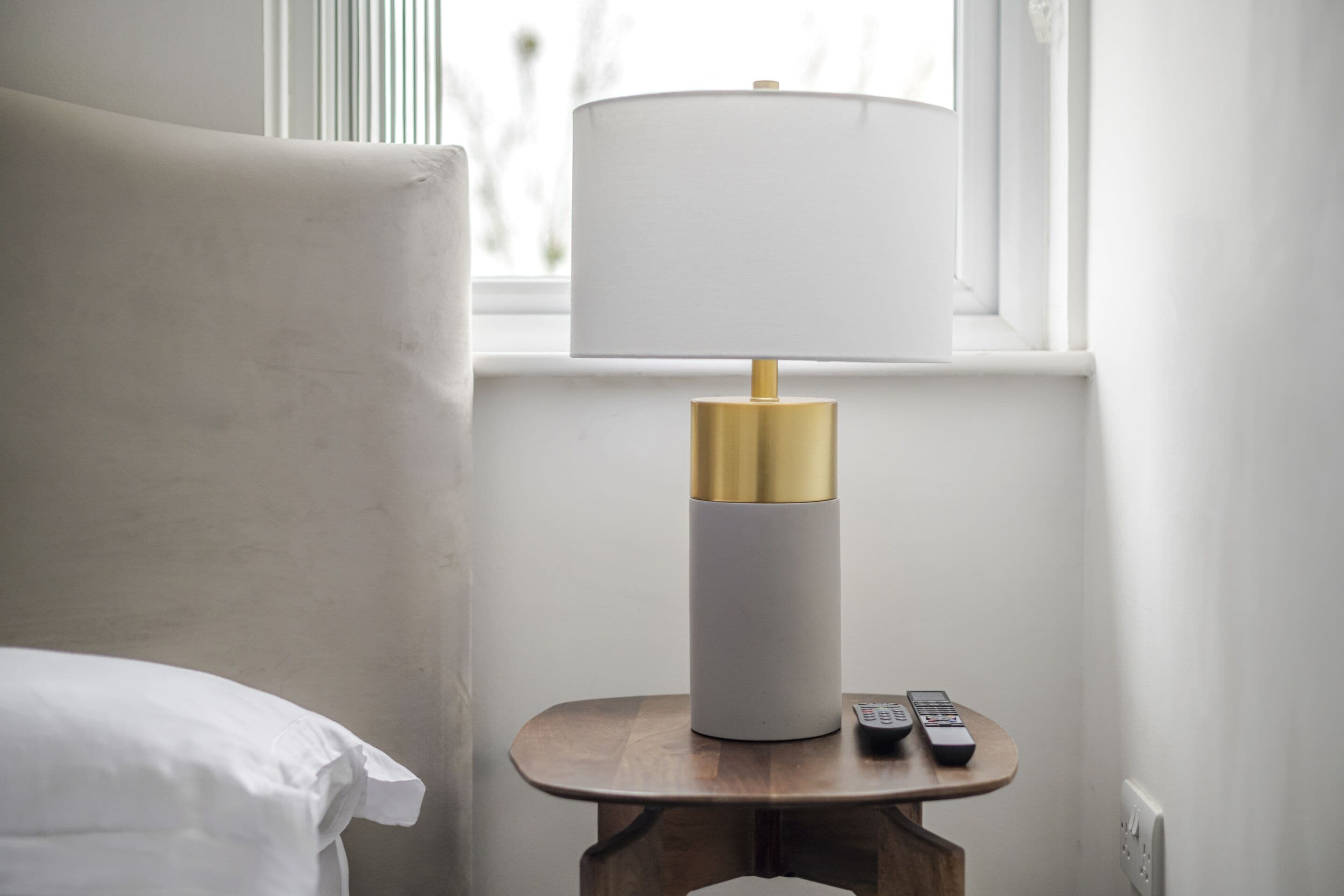 A bed with white bedding, a bedside table with a lamp, and two remote controls on the table.