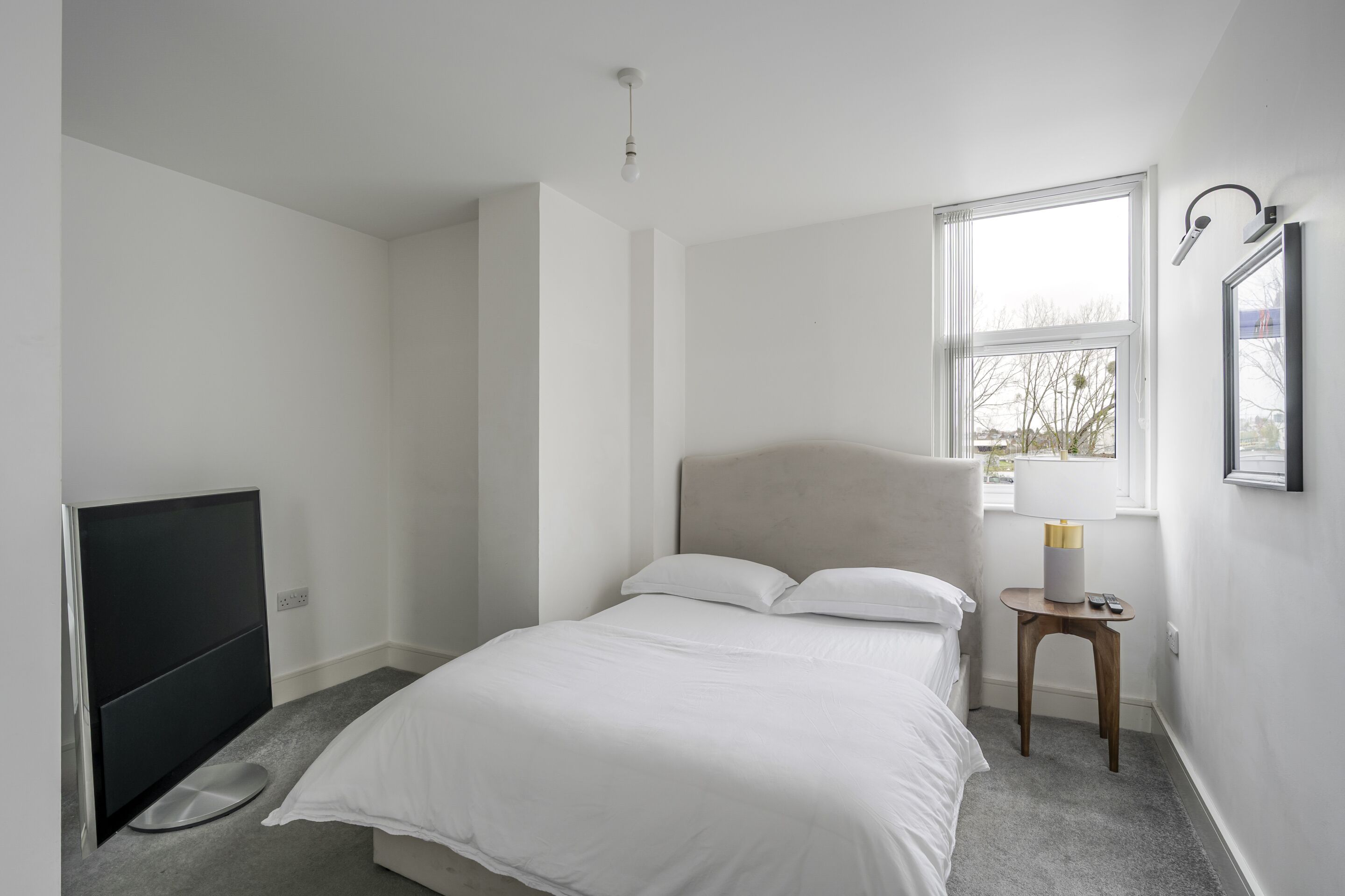 A bedroom with a bed, a TV, a window, and a bedside table with a lamp.