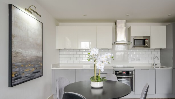 Cocina moderna con gabinetes blancos, microondas y estufa. Hay una mesa de comedor con sillas y un florero con flores blancas.