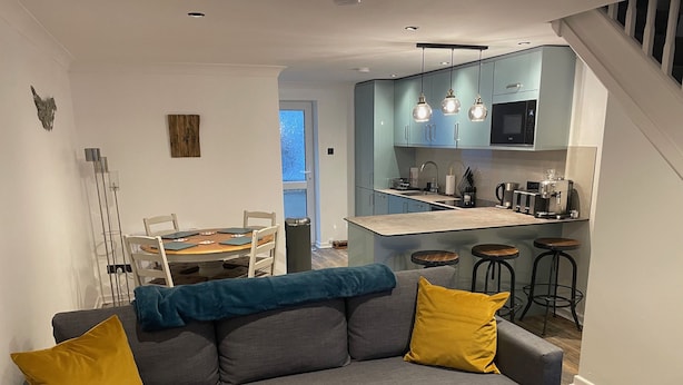 A modern living room with a grey sofa, two yellow cushions, and a blue throw blanket. A dining table with chairs is visible in the background. - 51 Fernhill Heights (Bridport)