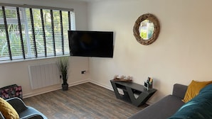 A modern living room with a flat-screen TV mounted on the wall, a dark console table with decorative items, a grey sofa with yellow cushions, a black armchair, and a potted plant. - 51 Fernhill Heights (Bridport)