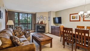 A living room with a sofa, coffee table, TV, fireplace, and dining area. - Pinnacle F01 Condo | Pool, Hot Tub | Golf Nearby (Killington)
