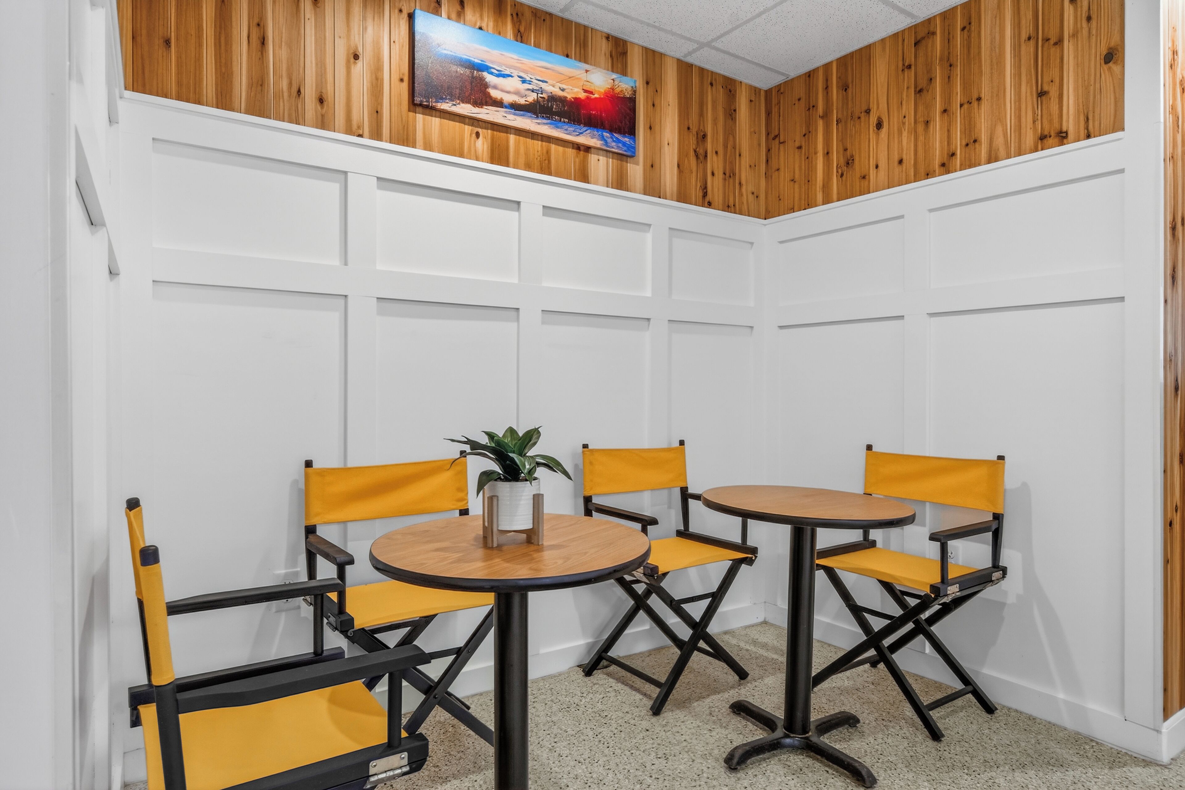 A corner room with white paneled walls, a wooden ceiling, and a painting. There are two round tables with yellow chairs and a potted plant on one table.