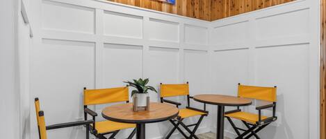 A corner room with white paneled walls, a wooden ceiling, and a painting. There are two round tables with yellow chairs and a potted plant on one table.