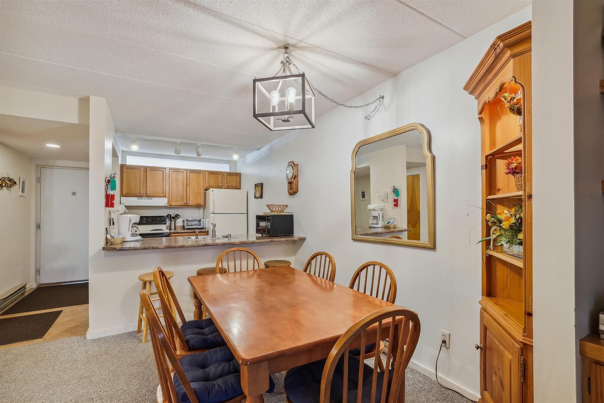 A dining area with a wooden table and chairs, a kitchen in the background, and a large mirror on the wall.