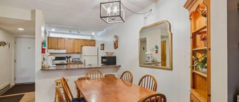 A dining area with a wooden table and chairs, a kitchen in the background, and a large mirror on the wall.