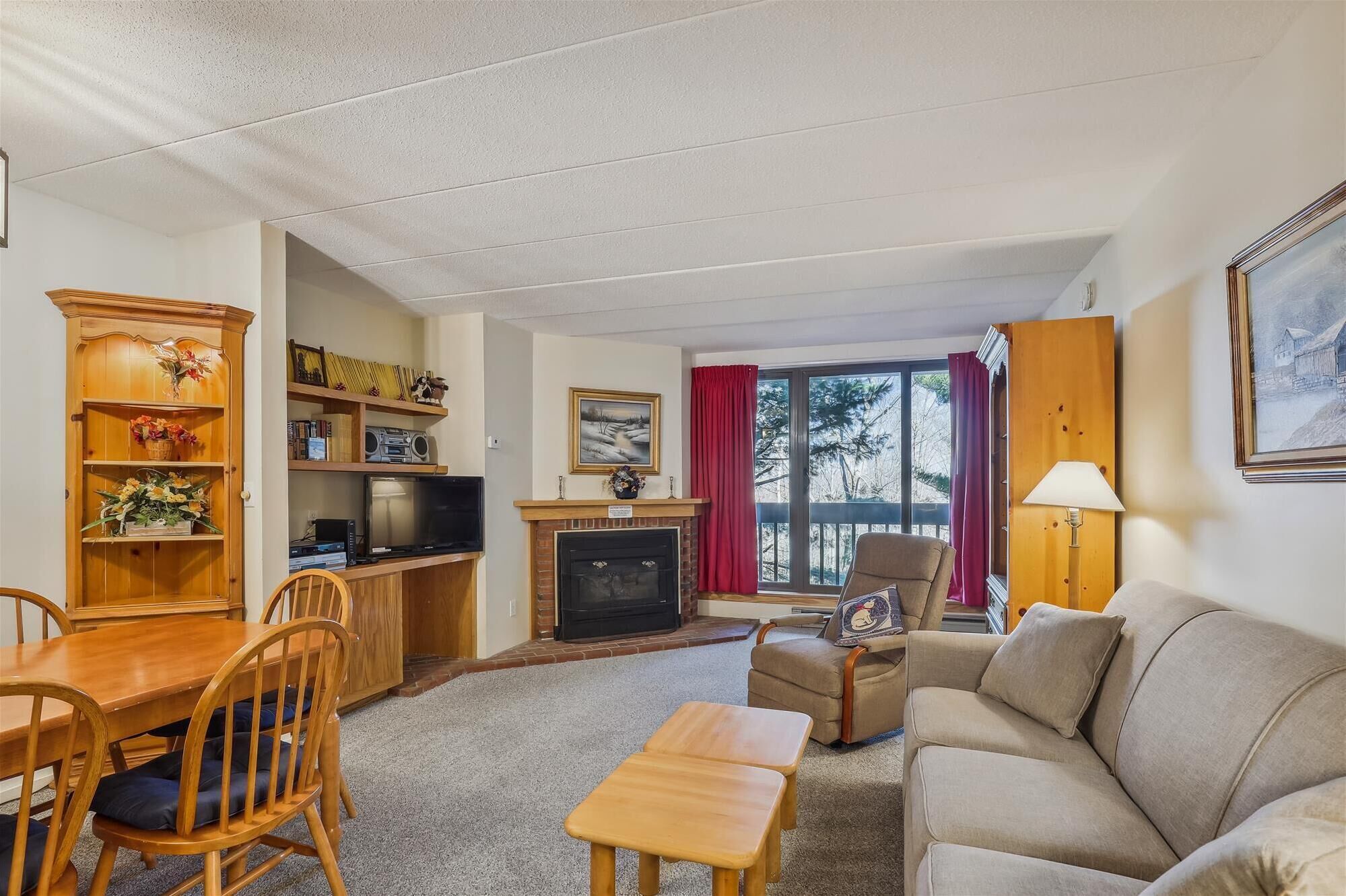 A cozy living room with a fireplace, a dining area with wooden furniture, and a large window with a view of trees.