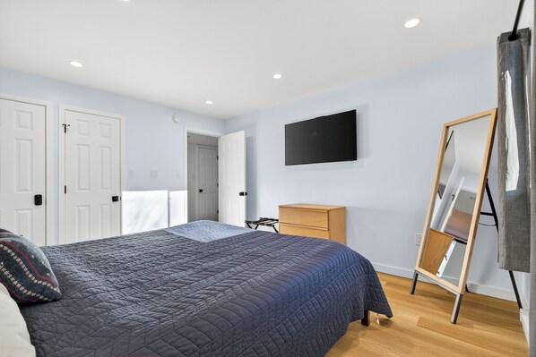 Een slaapkamer met een groot bed, een houten dressoir, een aan de muur bevestigde televisie en een spiegel die tegen de muur staat.