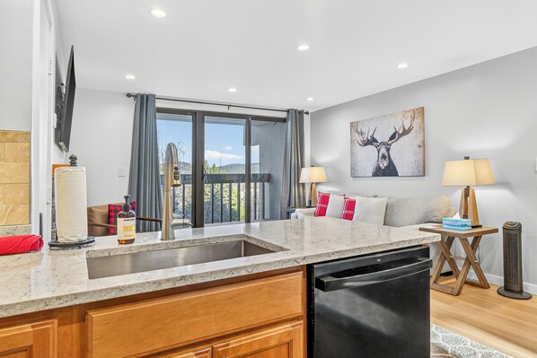 A modern kitchen with a built-in dishwasher, a large window with curtains, and a dining area with a table and chairs. - Pinnacle A13 Condo | Pool, Hot Tub | Golf Nearby (Killington)