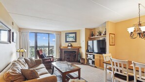 A living room with a sofa, coffee table, dining table, chairs, a fireplace, a TV, and a large window. - Pinnacle Condo | Pool, Hot Tub | Golf Nearby A15 (Killington)