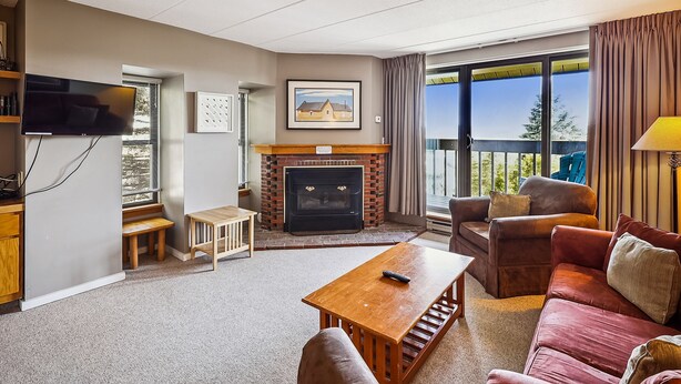 A living room with a fireplace, a sofa, a table, and a TV. - Pinnacle Condo | Pool, Hot Tub | Golf Nearby A17 (Killington)