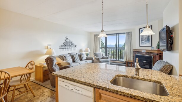 A modern kitchen with granite countertops, a dishwasher, and a sink. There is a dining area with a wooden table and chairs. The kitchen is adjacent to a living area with a sofa and a fireplace.