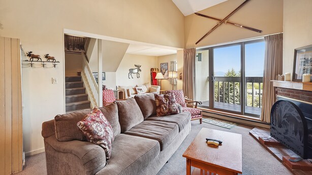 Ein gemütliches Wohnzimmer mit einer Couch, einem Holztisch und einem Kamin. Eine Treppe führt zu einer weiteren Ebene, und ein großes Fenster bietet Ausblick ins Freie.