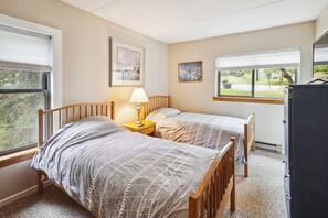 A bedroom with two beds, a nightstand with a lamp, a dresser, and a window with a view of greenery. - Pinnacle F08  Condo | Pool, Hot Tub | Golf Nearby (Killington)