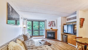 A living room with a fireplace, a television, and a large window overlooking a deck. - Pinnacle B15 Condo | Pool, Hot Tub | Golf Nearby (Killington)