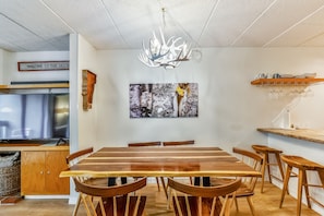 A dining area with a wooden table and chairs, a chandelier, a painting, and a television in the background.