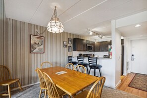 A dining area with a wooden table and chairs, a hanging light fixture, and a kitchen in the background.