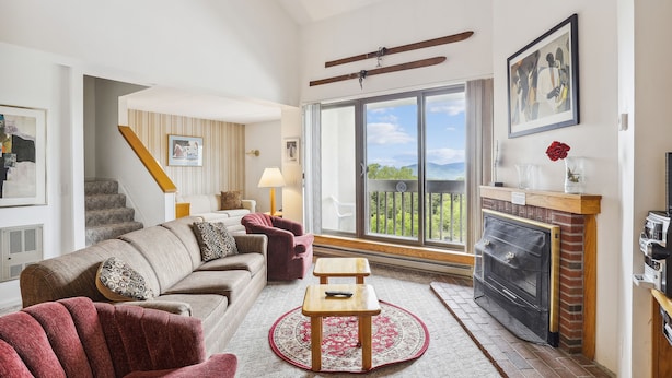 A spacious living room with a fireplace, a large window with a view, a staircase, and a cozy seating area. - Pinnacle C25 Condo | Pool, Hot Tub | Golf Nearby (Killington)