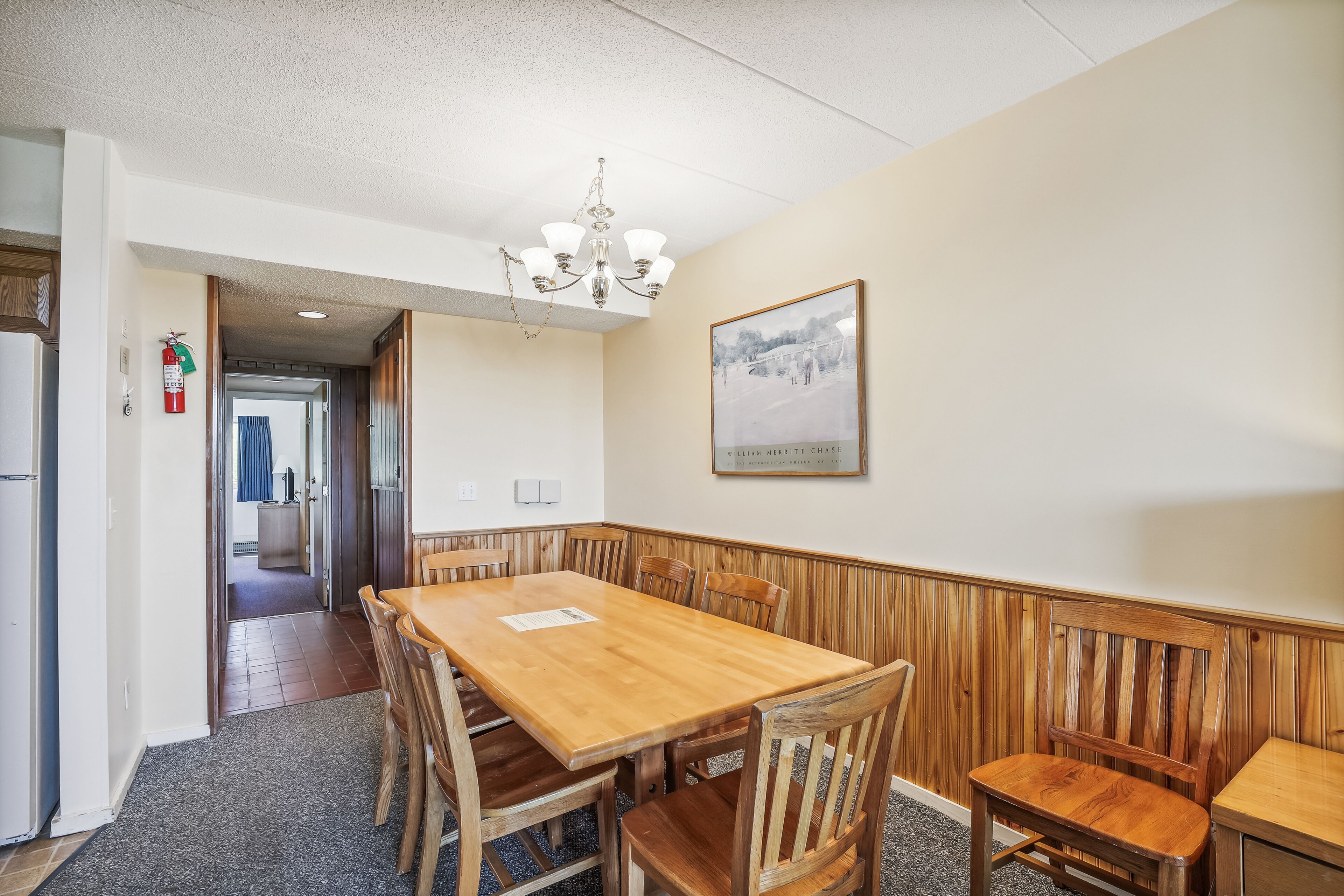 A dining area with a wooden table and chairs, a chandelier, a framed picture on the wall, and a fire extinguisher.