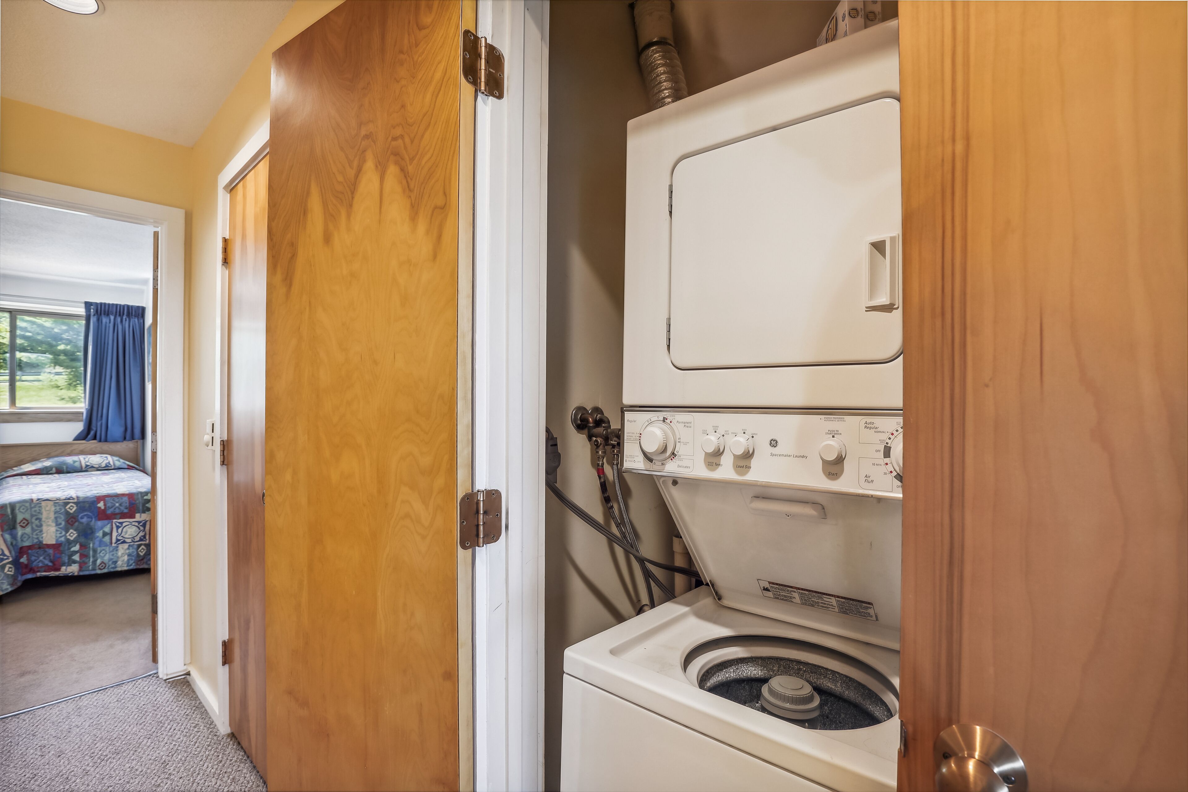 Une buanderie équipée d’un lave-linge et d’un sèche-linge à chargement frontal, de portes en bois, et une chambre adjacente avec un lit recouvert d’une couvre-lit à motifs.