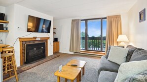 Uma sala de estar com lareira, televisão, sofá, mesa de centro e varanda com cortinas.