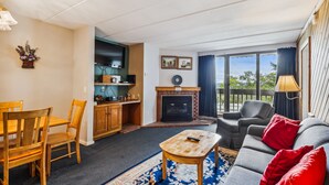 A living room with a fireplace, a dining area with wooden furniture, and a balcony with a view. - Pinnacle C09 Condo | Pool, Hot Tub | Golf Nearby (Killington)