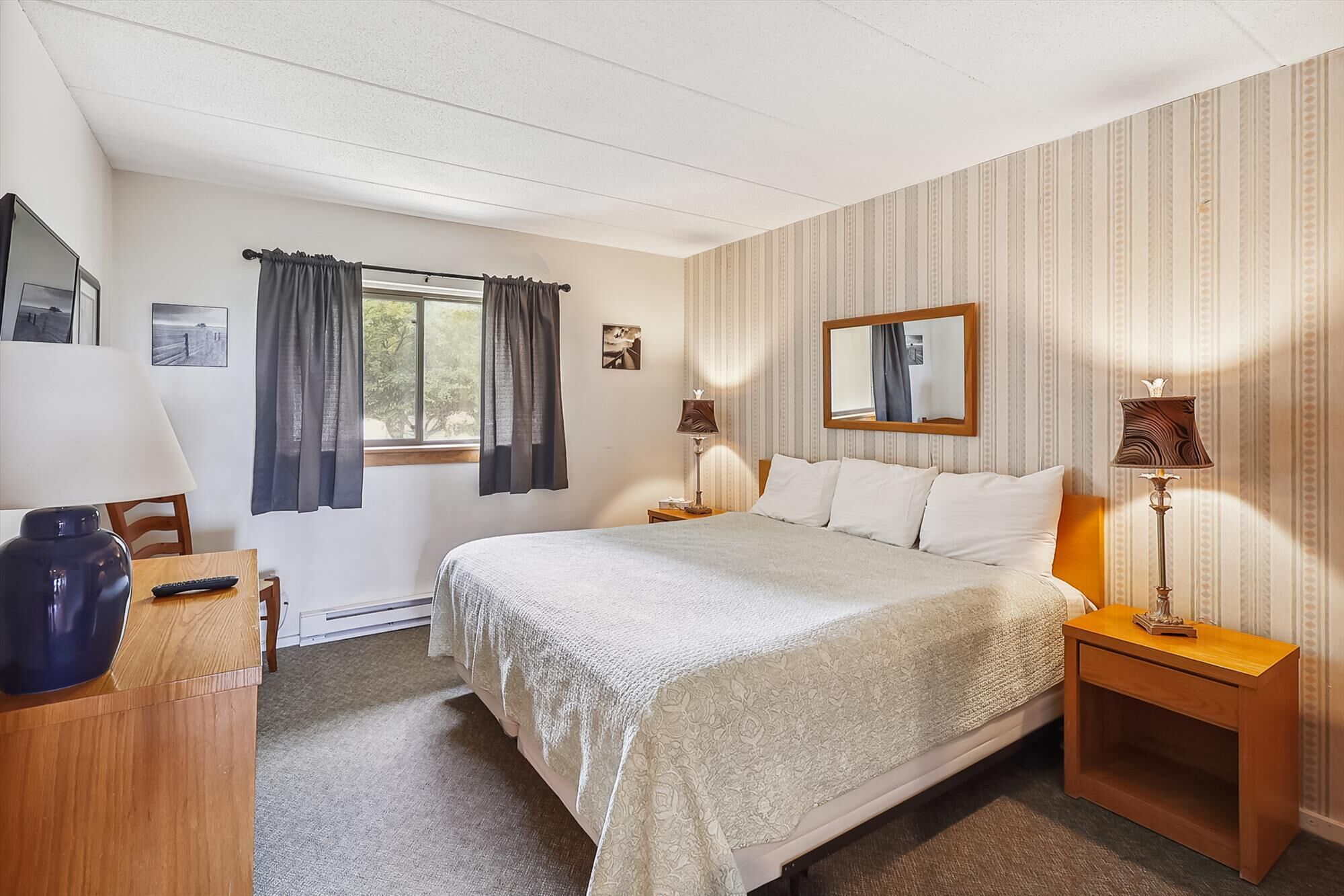 A bedroom with a bed, two bedside tables, a desk, a chair, a window with curtains, and a wall with a mirror and pictures.