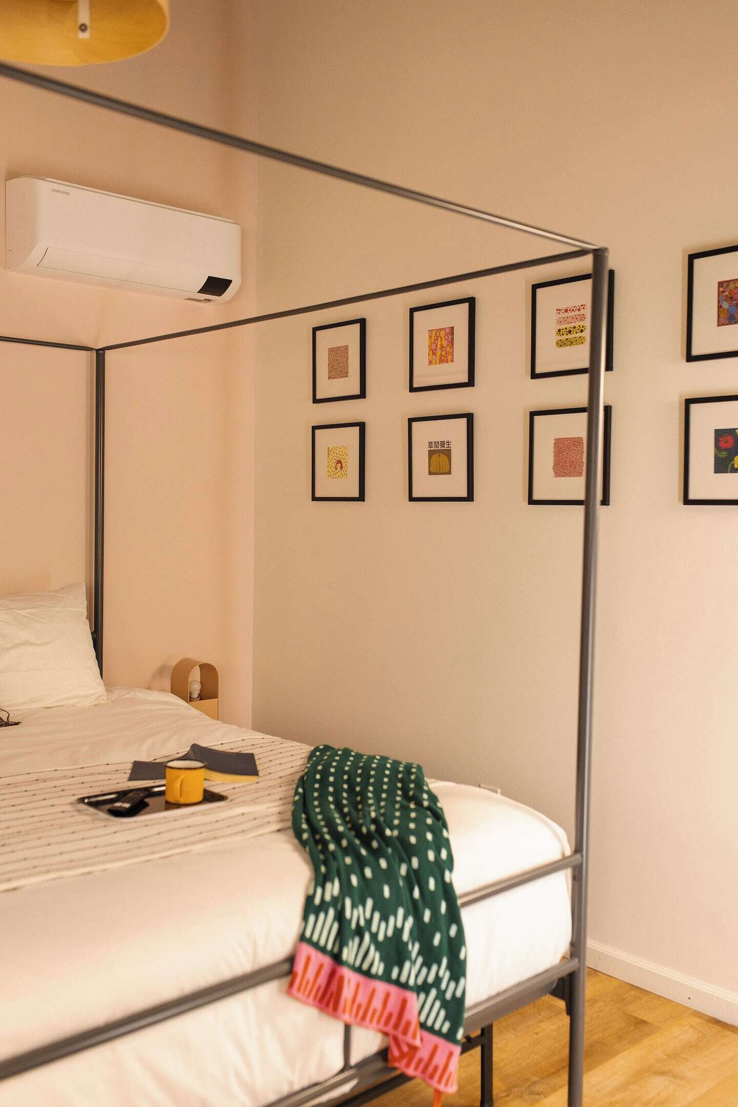 En säng med en metallram, en vit madrass och ett grönt, mönstrat överkast. På väggen hänger inramade bilder, och det står ett litet bord med ett gult föremål på.