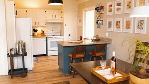 Une cuisine moderne avec des meubles blancs, un îlot central doté de tabourets de bar, un luminaire suspendu et un mur orné d’œuvres d’art encadrées.