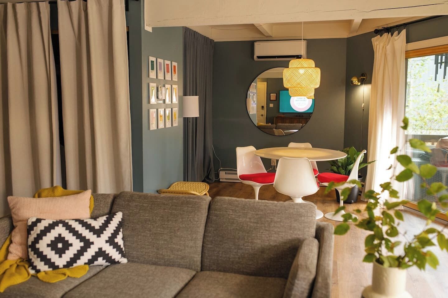 En stue med en grå sofa, et rundt bord med stole og et spejl på væggen.