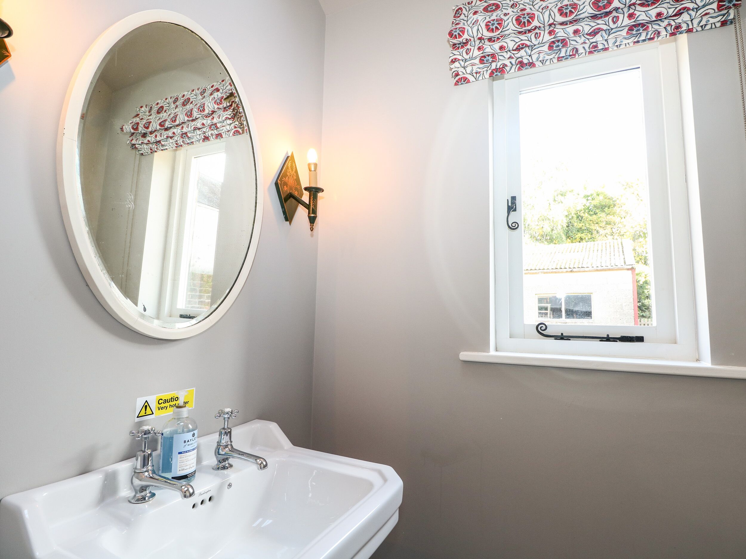 Een badkamer met een witte wastafel, een ronde spiegel, een raam met bloemen gordijn en een wandlamp.