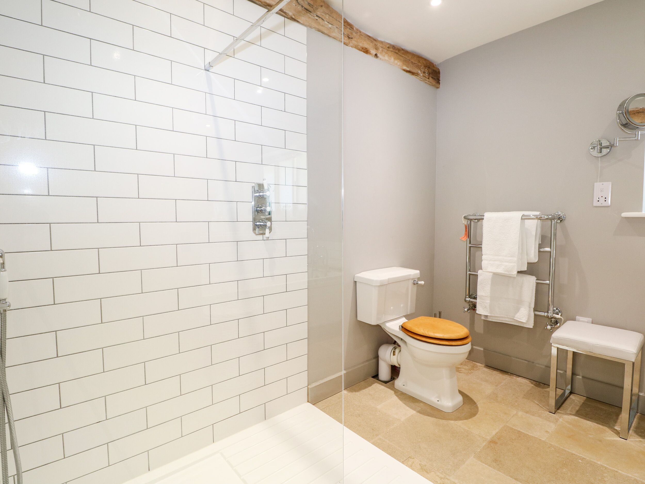 Un bagno con piastrelle bianche a metro, un soffitto con travi in legno, un water con seduta in legno, una panca bianca e un porta asciugamani.