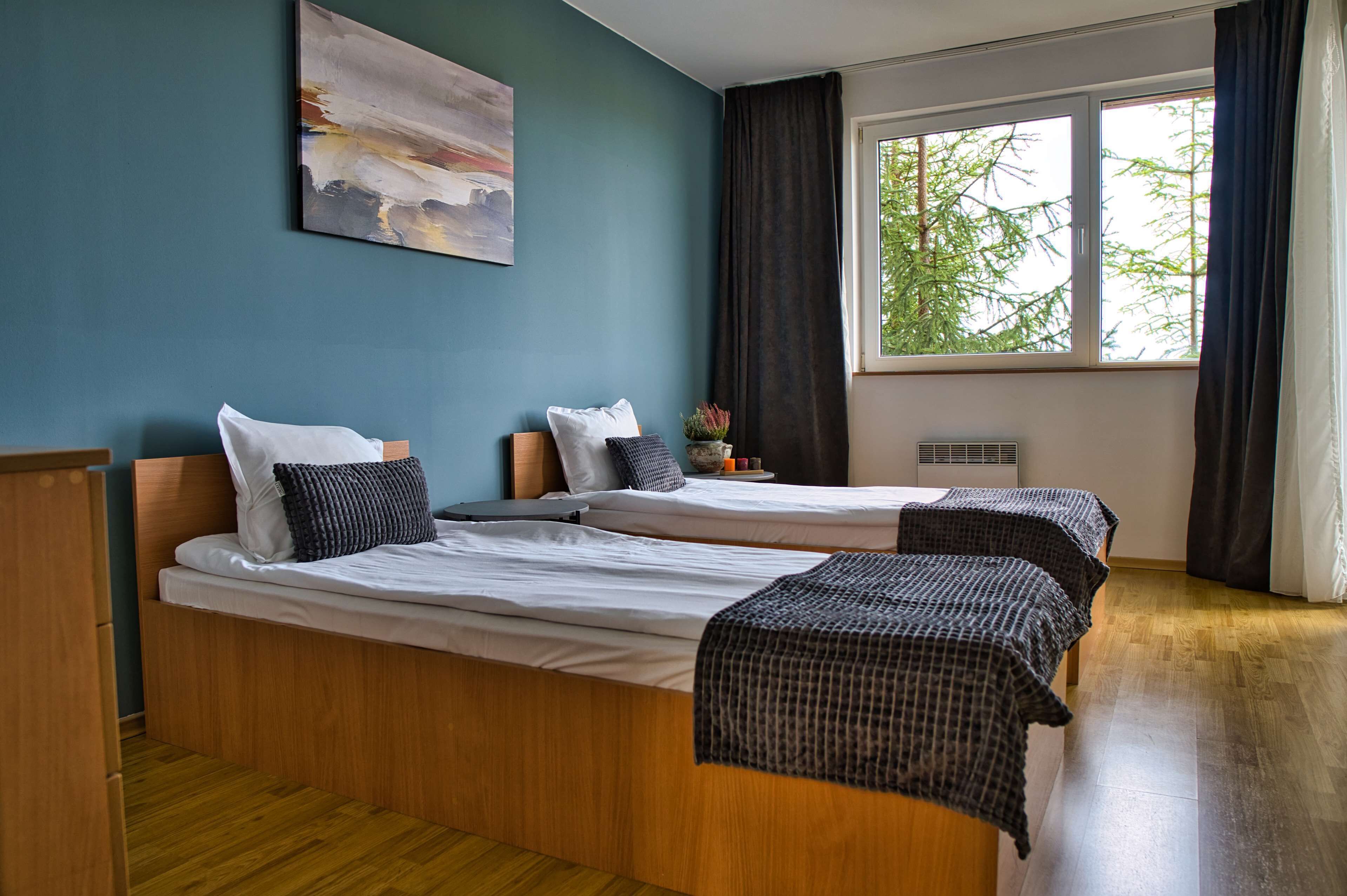 A room with two beds, a window with curtains, a painting on the wall, and a small table with a plant.