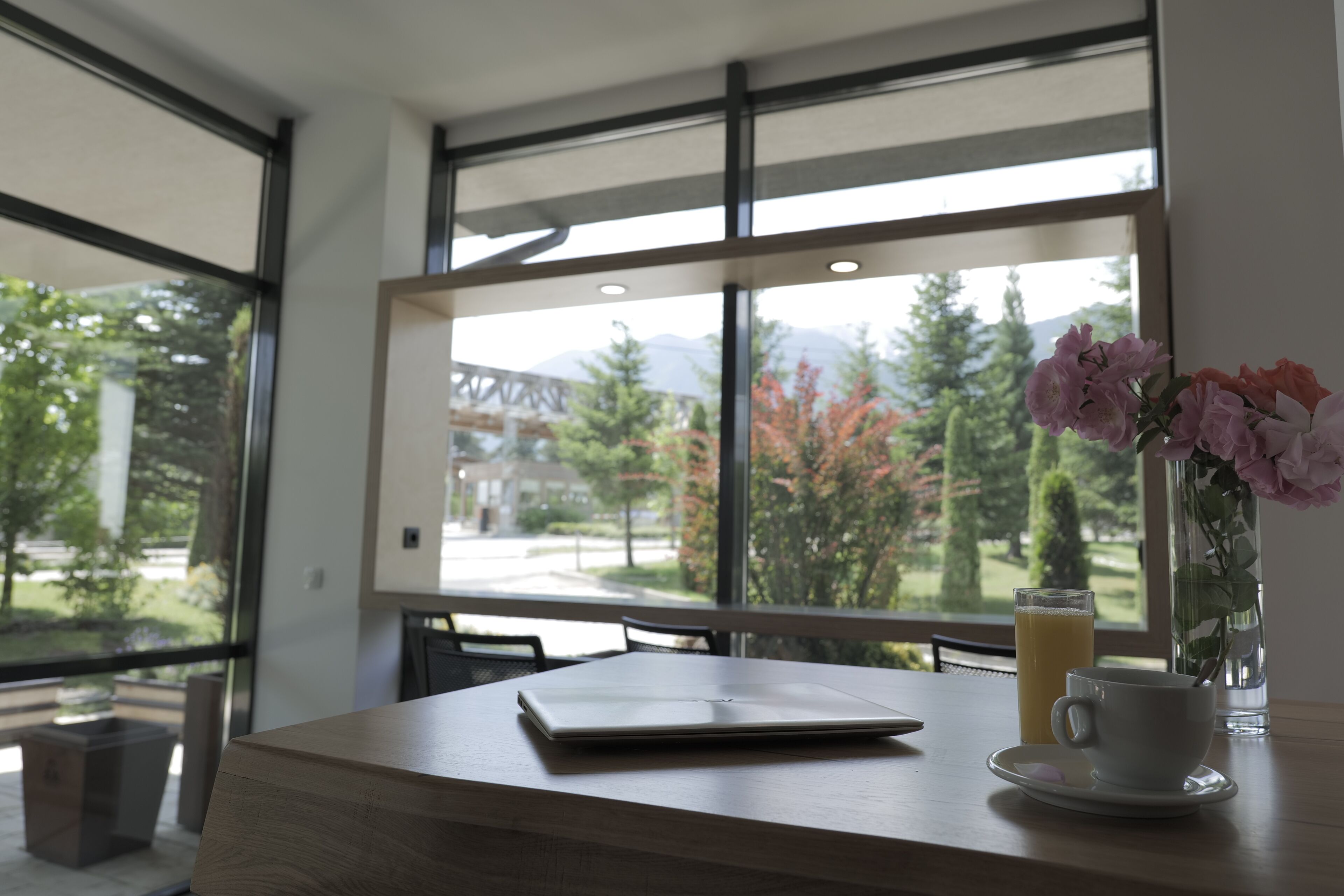 Une chambre moderne avec une grande fenêtre, un ordinateur portable posé sur une table, une tasse de café et un vase rempli de fleurs.