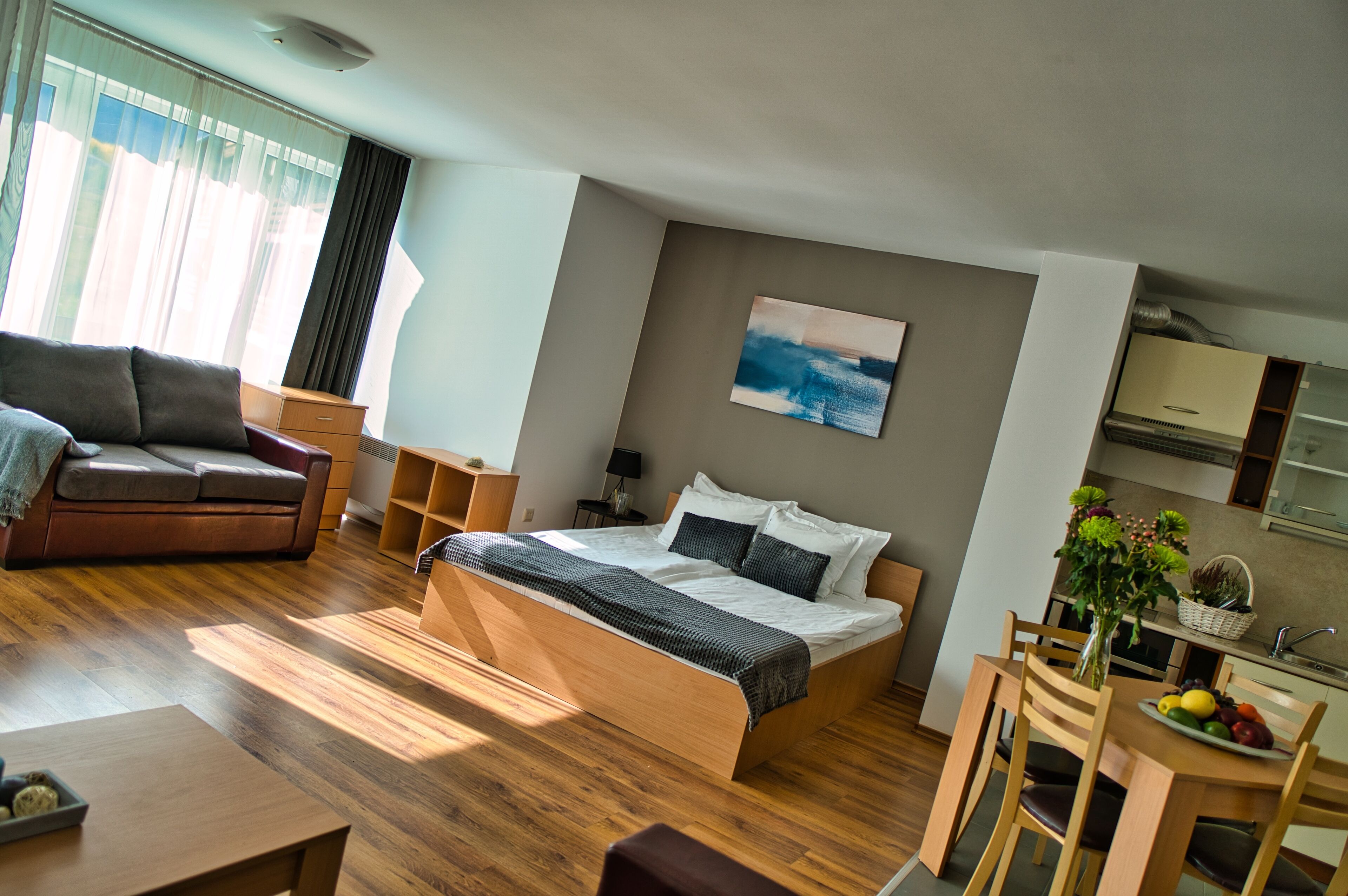 A modern bedroom with a wooden bed, a sofa, a dining table with chairs, and a kitchen area.