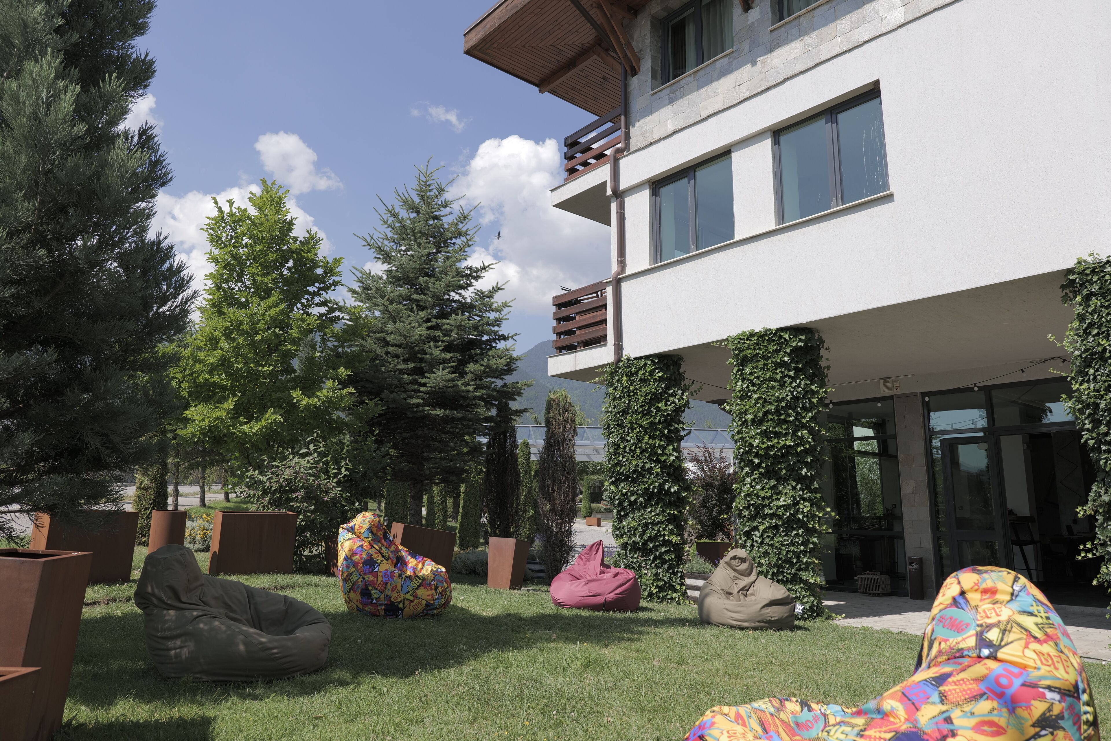 Un edificio moderno con techo plano, ventanas amplias y un balcón. Se observan varios pufs de colores y un área verde al frente.