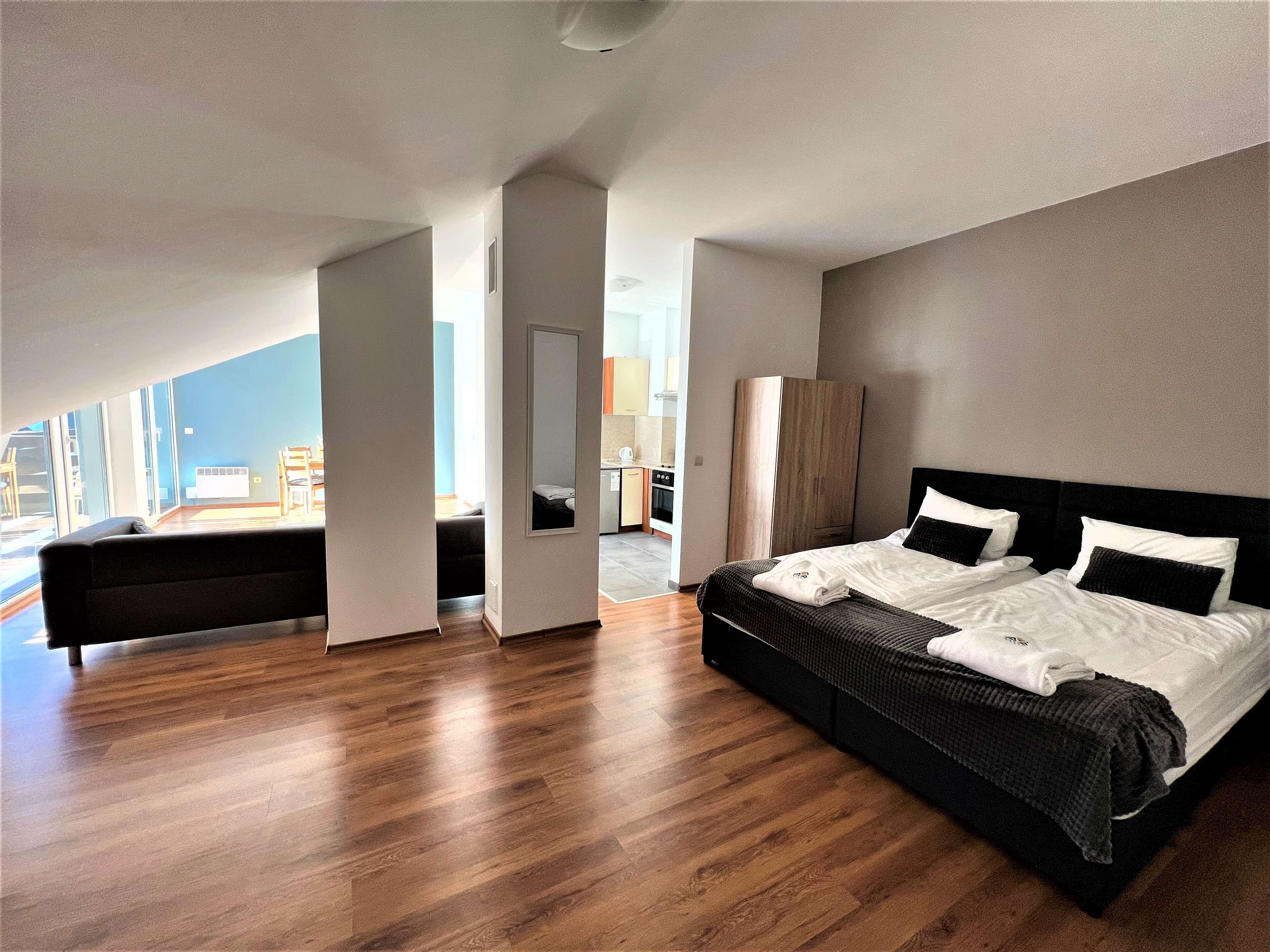 A modern hotel room with a large bed, a sofa, and a view of the kitchen and dining area.