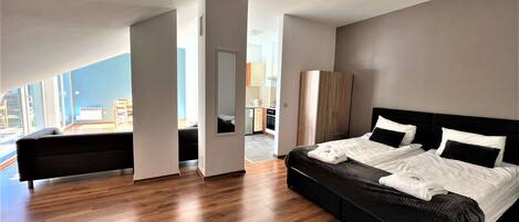 A modern hotel room with a large bed, a sofa, and a view of the kitchen and dining area.