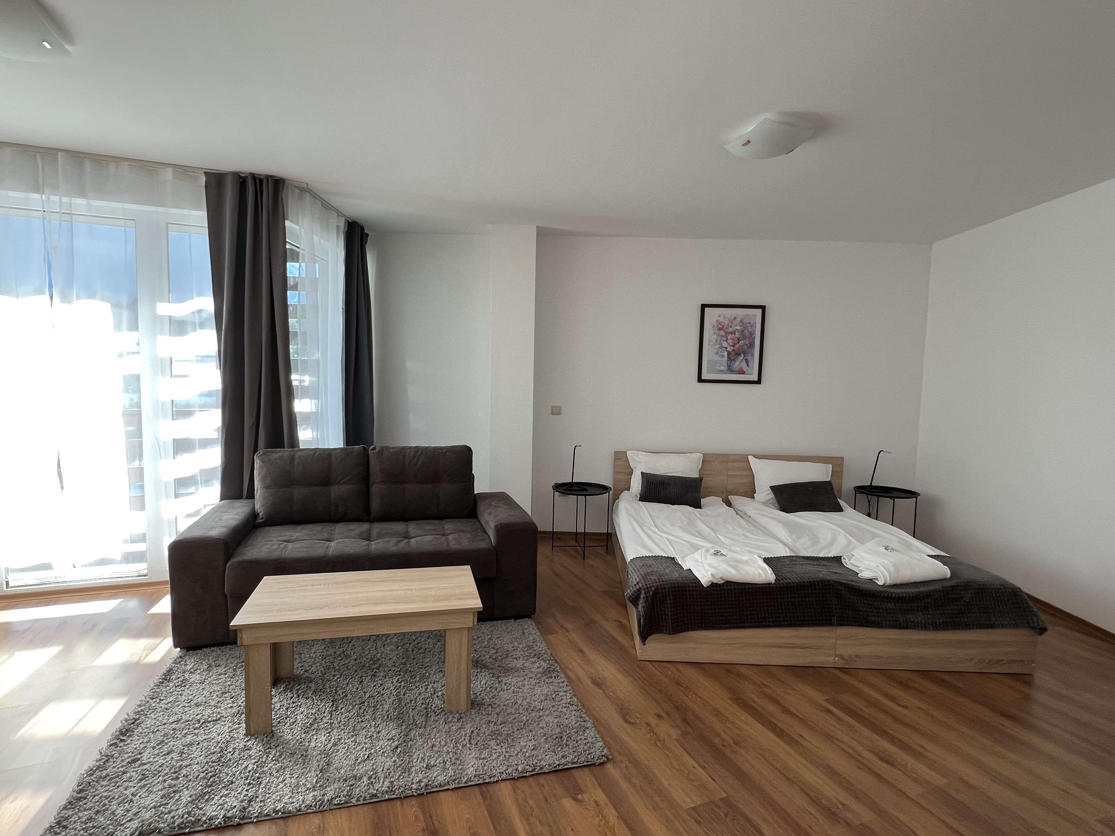 A modern bedroom with a sofa, a bed, a small table, and a view of the city through large windows.