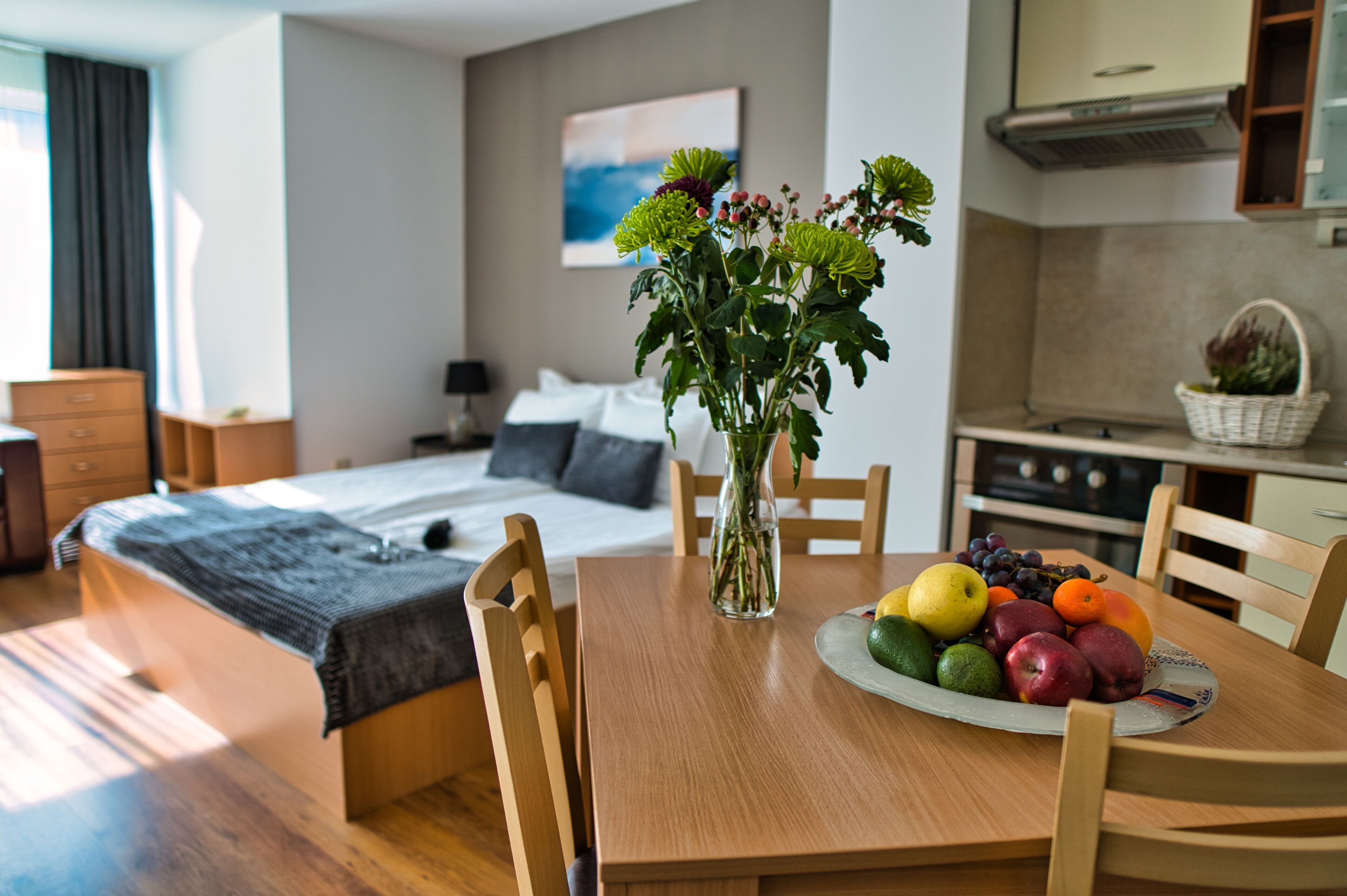 A modern hotel room with a wooden bed, a dining table with fruit, and a vase of flowers.